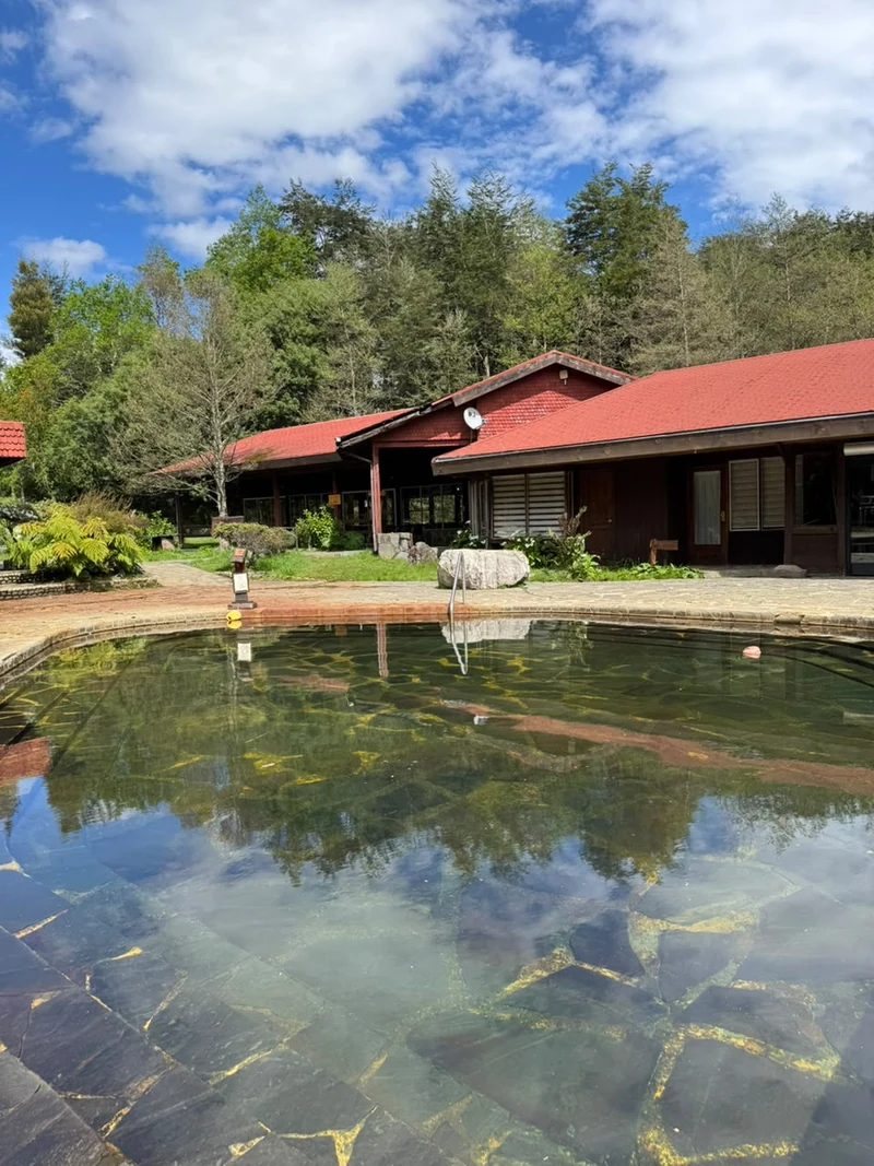 Piscina exterior de Termas Coñaripe junto a construcciones de techo rojo y bosque
